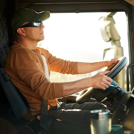 Person seated in the driver’s seat of a large vehicle, holding the steering wheel with both hands. The individual is wearing a brown long-sleeve shirt and a green cap