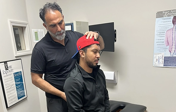 Dr. Karm Virk performing a neck adjustment on a seated patient wearing a red cap in a clinical setting, with medical posters and equipment visible in the background