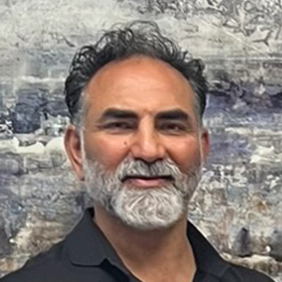 Dr. Karm Virk wearing a black collared shirt standing in front of a textured gray and white wall